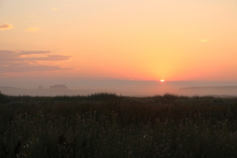 The sun peeks over the horizon at Agate Fossil Beds National Monument. The visitors center and the sun are partially obscured by fog. It is 6:17 a.m.